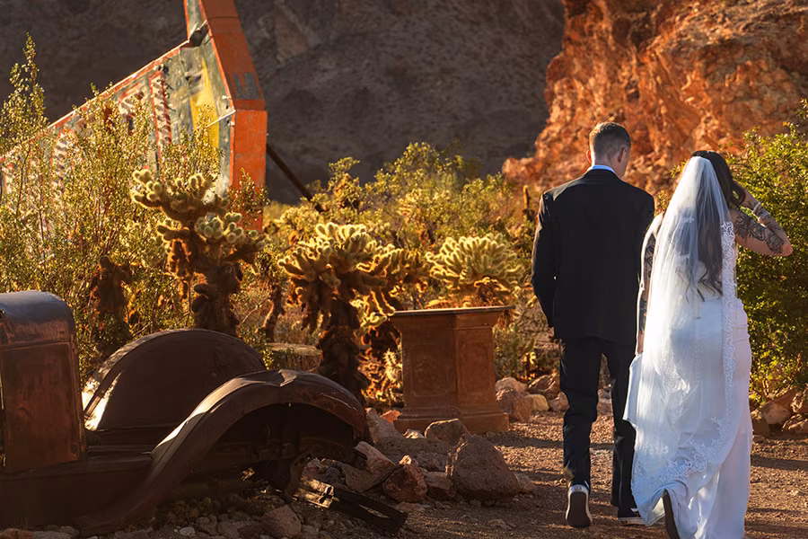 Bride and groom walking past vintage rusted trucks and cholla cactus at Nelson Ghost Town