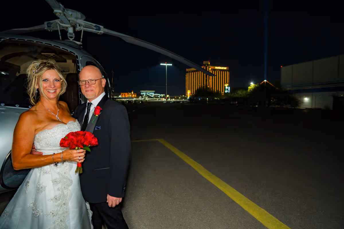 Bride and groom with red roses beside their helicopter at the Las Vegas helipad, Mandalay Bay lit up behind them