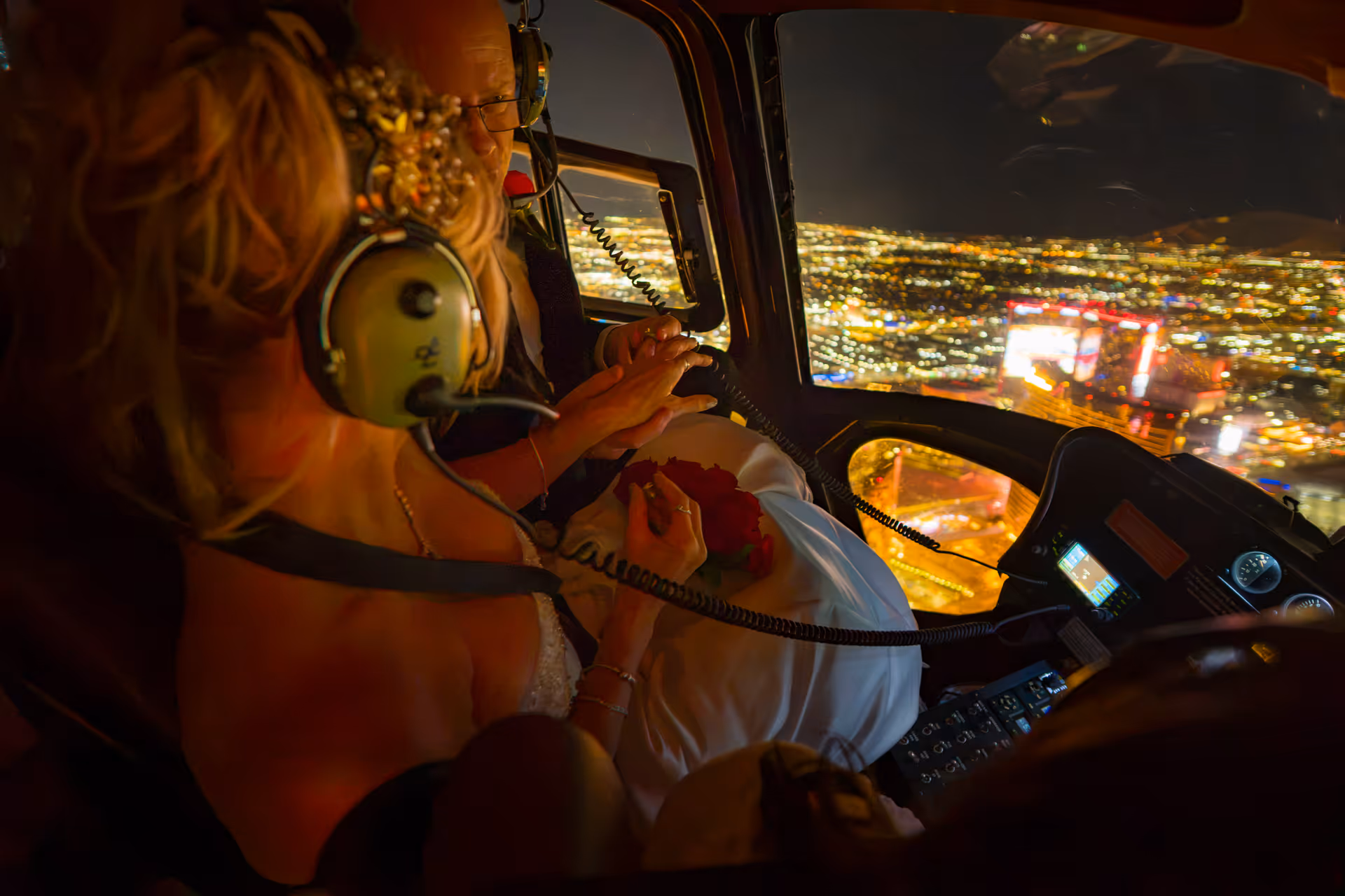 Bride and groom holding hands with red roses inside a helicopter over the Las Vegas Strip lights at night
